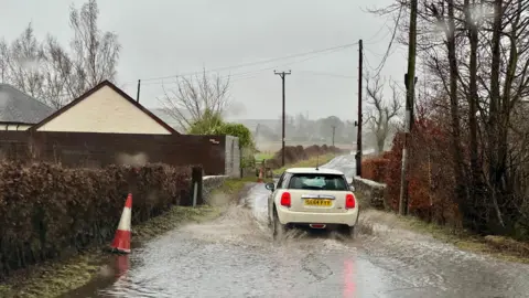 Nellington/BBC Weather Watchers A white mini makes its way through water on a narrow rural road with a house to the left and trees to the right