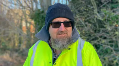 Laura Coffey/BBC Man with black hat and beard wearing a high-vis jacket and sunglasses, and a blue beanie hat. There are trees in the background