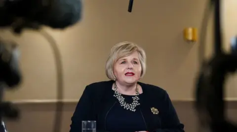 PA Media Donna Ockenden seated at a desk at a press conference. He wears black and has a big necklace. The background is slightly blurred, but the cameras can be seen. 