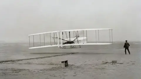 Getty Images A black and white image of a plane used by the Wright brothers. A man stands to the left of the plane, with his back facing the camera.