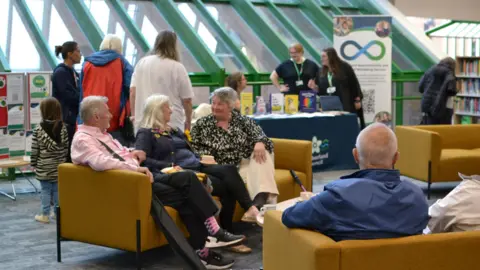 A man and two women are talking to each other as they sit on a mustard-colour sofa at Carlisle Library. A young girl is standing behind them looking at a display board. In the background, some books are positioned on a table as three members of library staff chat.