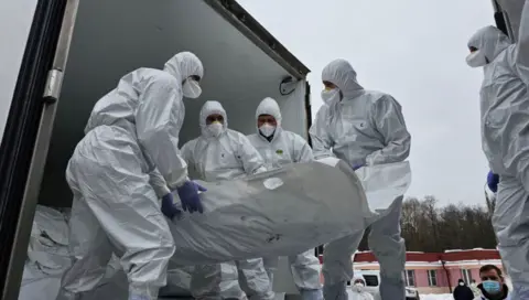 Officials in white overalls that cover their heads and wearing face mask carry a white stretcher presumably containing a body being unloaded from what looks like a truck