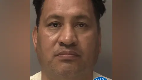 A police mugshot of a man with dark hair. He is wearing a cream jumper. There is a West Midlands Police logo in the bottom right corner.