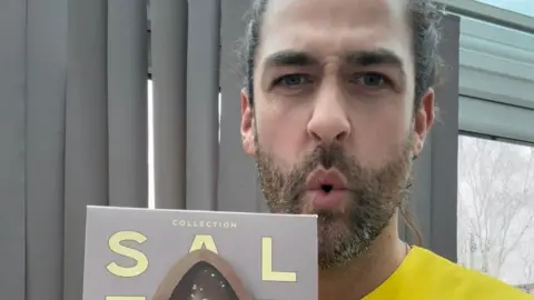 Billy D'Souza A man with his hair tied up with a puckered mouth as he holds a packaged Easter egg