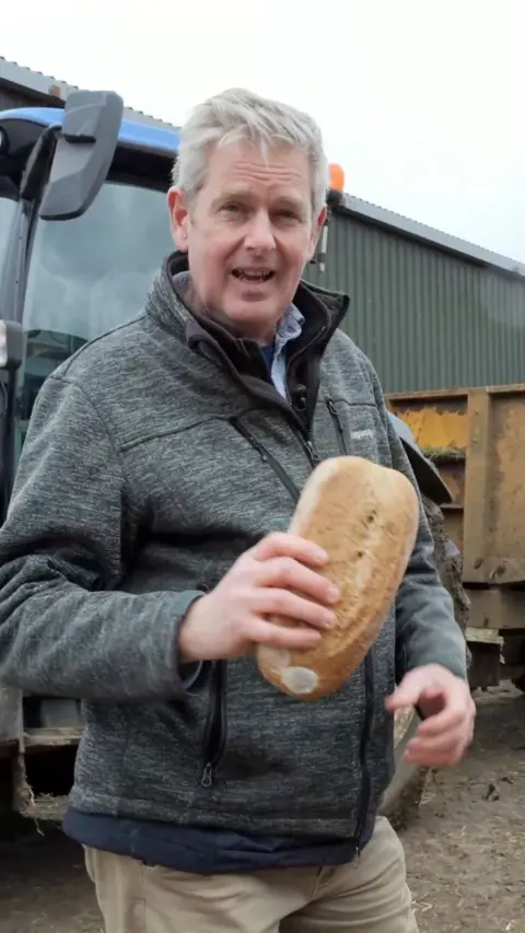 A man holding a baguette whilst standing in front of a tractor on a farm.