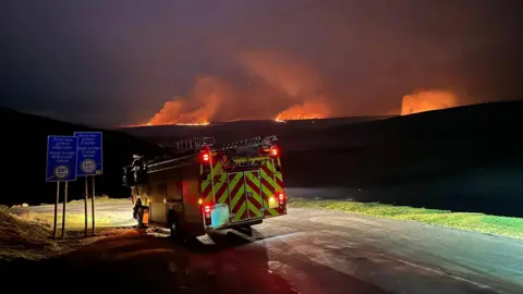 MWWFRS Rhayader A fire engine is pictured on a road under the night sky. Wildfires have lit up the dark with several spots pictured wit  large amounts of smoke.