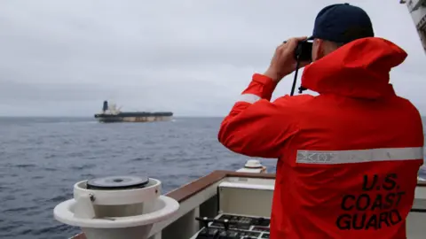 A US Coast Guard officer wearing a cap and a bright red jacket uses a pair of binoculars to look at the tanker.