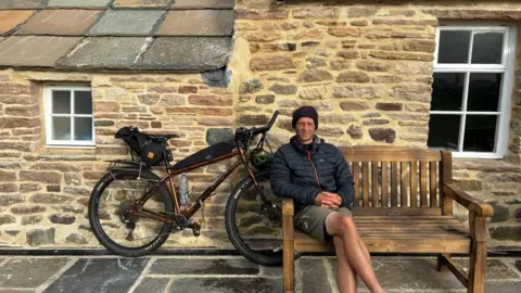 A man sits on a bench outside Burnmouth bothy. He is wearing a jacket, shorts, and a headband. Behind him is the bike that has joined him for the last five years on a world tour.