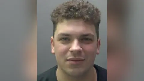 A police image of Jake Cummings: The head and shoulders of a man with dark, curly, hair wearing a black, round-necked top.