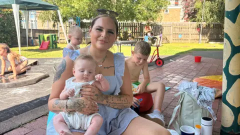 A mother wearing a dress is smiling for a photo while holding her baby boy. Other children are seen playing in the yard behind her. There is a bag and other personal items nearby. It is sunny.
