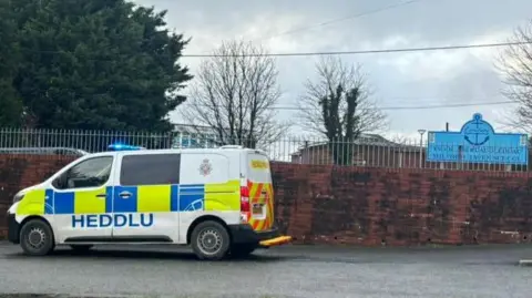 A brown school wall with a silver railing above and a police van parked outside. In the background is a blue school sign.