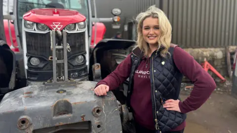 Grace is next to a tractor, she uses a Burgundy sweater under a blue padded gillet. She has wavy blond hair to her shoulders and smiles at the camera.