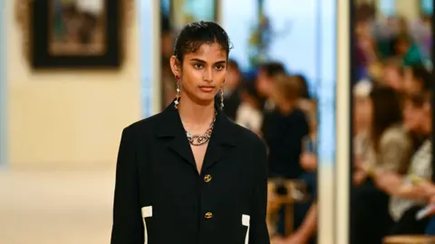 AFP via Getty Images Indian fashion model Bhavitha Mandava presents a creation for the Chanel Cruise 2026/2027 collection in Biarritz on April 28, 2026. She's wearing a black blazer with white detailing on the sides and golden buttons. 