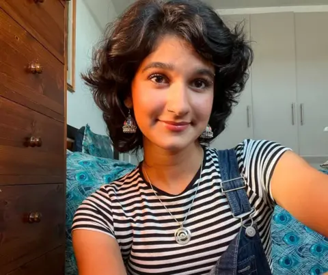 Fatima Qureshi A woman in a striped black and white T-shirt sits in a bedroom and smiles at the camera