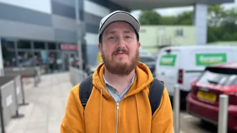 A man wearing a yellow hoodie and a black backpack. He has ginger hair and a white cap on. In the background there are cars and a retain park including a closed cineworld.