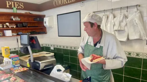 A man dress in white overalls with a green and white striped apron holding a  rare steak in a piece of paper. He is looking away from the camera towards a till and a Paxo box can be seen on the counter next to a yellow charity donation collection box