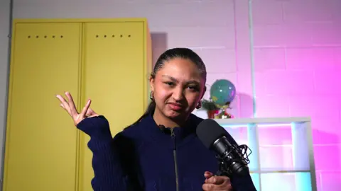 A young woman wearing a dark zip‑up sweater speaks into a studio microphone, in a softly lit background.
