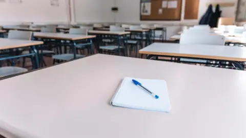 A general view of a generic classroom. A notebook with a pen on a table is in the centre of the picture, with desks behind it.