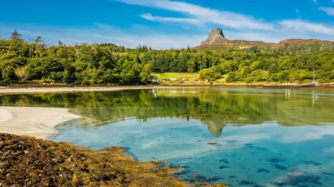 Getty Images A scene on Eigg with a beach, and mirror-like calm sea. On the opposite shore is an area of trees and its landmark hill, The Sgùrr.
