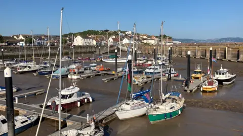 Watchet Harbour at low tide, with around 50 boats inside, with the town's esplanade in the background