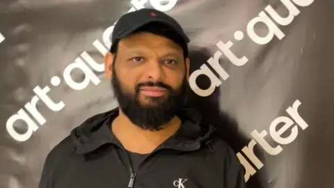 Jordan Patel, a man wearing a black jacket, is standing against a black step and repeat board which has white lettering featuring the logo of the Art Quarter.