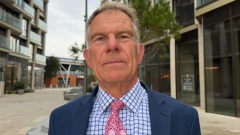 Christopher Schofield is pictured by high rise flats in St Helier, he is wearing a pink tie and a chequered shirt. 