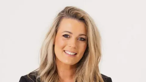 Lynn Wortley A woman with long blonde hair is smiling. She is wearing a black jacket and has a watch on her left wrist.