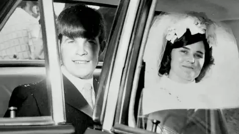 Family handout A black and white photo of Brian McCrory on his wedding day. He is sitting in the back of a car wearing a suit and tie beside his wife, who is wearing a white wedding dress and veil pulled back from her face.