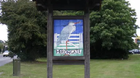 A village sign saying Herongate, with trees behind it and grass in front. The sign shows a heron with a blue sky. The sign is in a large wooden frame.
