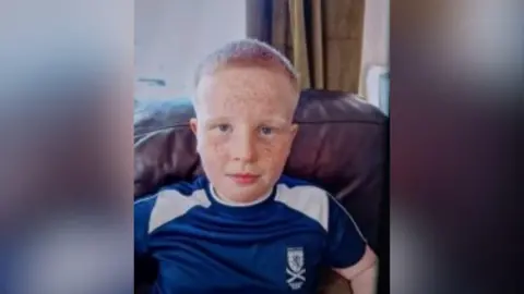 Haiden Crawford looking right at the camera. He has short, orange hair and dark eyes. He has freckles covering his facing. He is wearing a blue t shirt with a Scotland flag in the upper right corner.