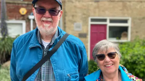 Alex Dunlop/BBC Ian is wearing a navy baseball hat, sunglasses and has a grey beard. He is wearing a denim jacket, checked shirt and has a bag strap across his body. Sara has grey short hair and is wearing a blue coat, with a colourful patterned hood, and a floral top. They are both smiling and standing in front of a building.