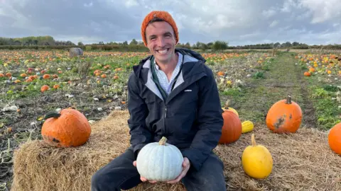 BBC Farmer George Holdstock sat on hay bale in pumpkin field holding grey pumpkin