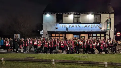 A crowd of people are wearing pink high-vis jackets, standing outside The Park Tavern pub. The building is painted white with a blue sign.