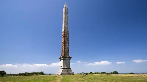 A wide shot of the Gilbert Memorial in front of a blue sky. The monument is very tall and weathered.