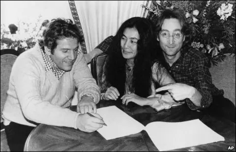 AP Allen Klein signs paperwork, with Yoko Ono and John Lennon sitting next to him at a small round table.  