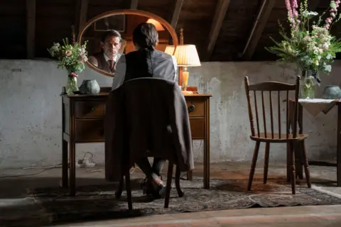 BBC/Mam Tor Productions/Anne Binckebanck A man in a suit, dressed for a wedding, sits looking at himself in a dressing table mirror.