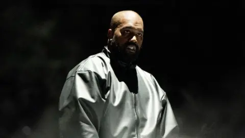 Getty Images Kanye West wears a black leather jacket and has a beam of light shining down upon him while he stands in front of a black background.