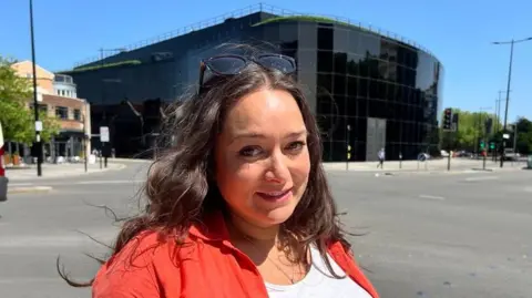 Stuart Howells/BBC Naomi Drown stands outside the Willis building. She is smiling and has long wavy brown hair. She wears a red shirt with a white T-shirt underneath. Sunglasses rest on top of her head.