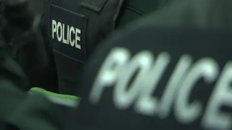 The picture shows a close-up of the back of police officers' black jackets with police in white letters 