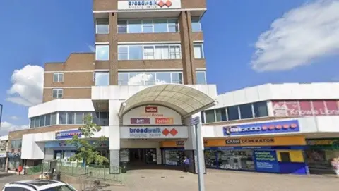 Google The exterior of the Broadwalk shopping centre, which is an old-fashioned 1960s-style brick building with a white awning over the front and the names of shops in coloured banners on the front