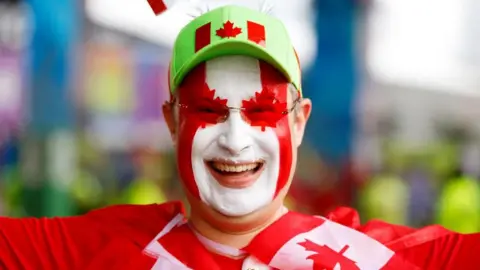 A fan smiles widely in red and white face paint, a red shirt, and a light green hat with a Canadian flag on the front a 