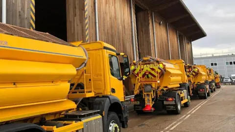 A fleet of yellow gritter lorries parked up in a row behind each other at a depot.