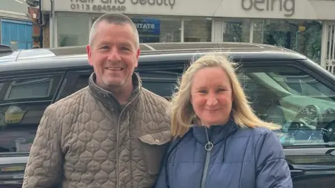 Grace Wood/BBC A man with a green quilted jacket and a woman in a blue puffer coat smile at the camera. They stand in front of a car on a busy road.