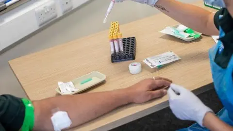 A medical worker wearing a blue PPE apron and white gloves sits at a desk after taking a blood sample from the arm of a patient, which is resting on the desk with a white swab on it. There is a tray of sample tubes on the table along with other medical equipment.