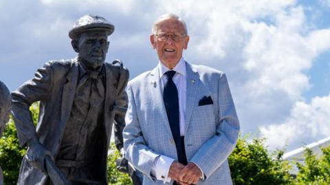 Johnny Ringwood standing next to the statue depicting himself, he is wearing a light blue suit jacket, a dark coloured tie and glasses