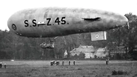 An airship floating above a field. Several small people can be seen on the ground underneath. The balloon has SSZ45 written on it.