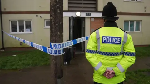 A police officer in a green hi-vis jacket stands in front of a house with his hands folded behind his back. In front of him is a house with blue and white police tape across part of the area. 