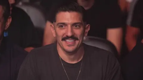 Getty Images Andrew Schulz, pictured in 2021 at a UFC event. Andrew has short dark hair, brown eyes, and a brown mustache. He wears a gold chain over a dark gray t-shirt and is pictured seated at the event, with the crowd behind him blurred. 