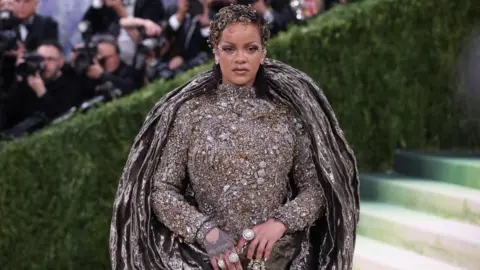 Rihanna wears a silver dress that wraps around her body, with large silver rings and her hair is adorned with gold jewellery. She is standing on steps with a number of photographers taking photos behind her. 