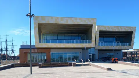 A modern-looking leisure centre with large windows and a large yellow tiled design. It is a sunny day. 